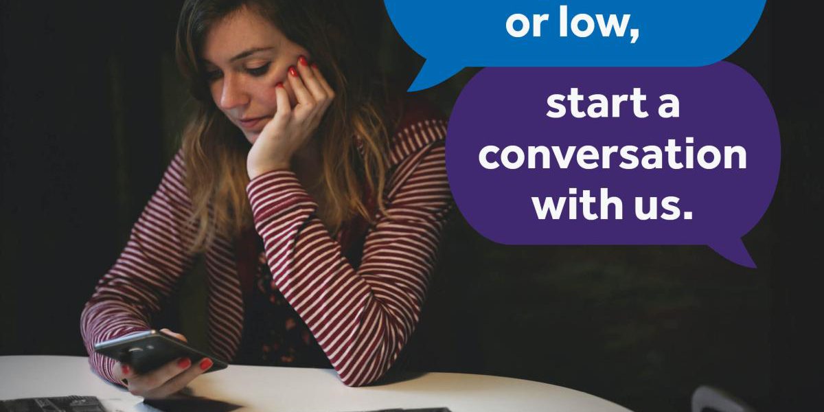 Woman using her phone in a dark room. Text bubbles say: If you're feeling anxious or low, start a conversation with us. White text on a blue banner reads: Text HEAR to 85258 Free confidential support 24/7. NHS Peterborough and Cambridgeshire, and in partnership with shout logos are on the bottom right corner.